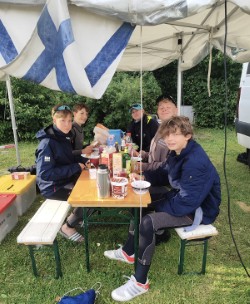 Seglerinnen und Segler beim gemeinsamen Essen an einer Bierzeltgarnitur unter einem Pavillon mit finnischer Flagge im WM-Camp in Schilksee