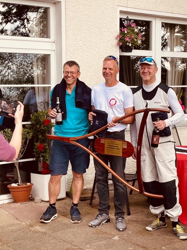 Drei O-Jollen-Segler bei der Siegerehrung des Champagner-Cups mit Sektflaschen und dem hölzernen Wanderpreis für Olympiajollen vor dem Clubhaus der Segler-Vereinigung Tegel