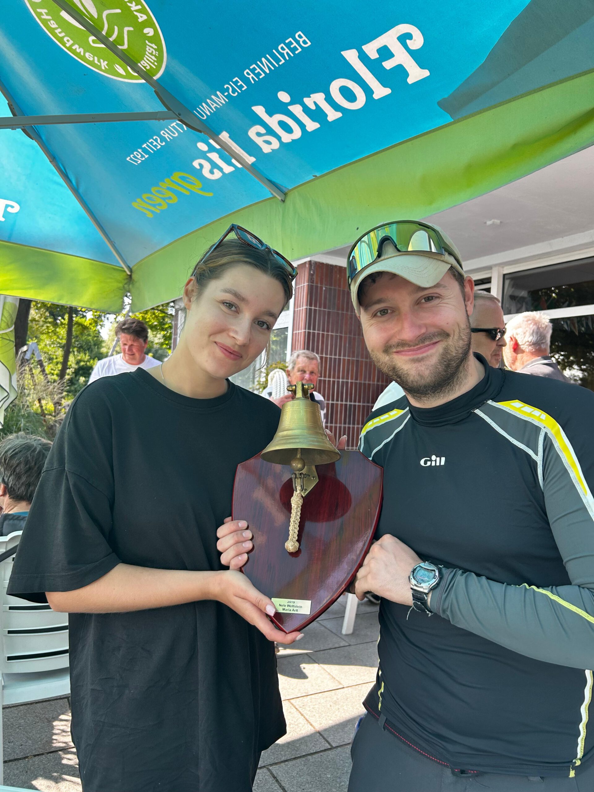 Siegerehrung des Ernstel-Cups: zwei Seglerinnen und Segler halten unter einem Sonnenschirm gemeinsam den hölzernen Wanderpreis mit Messingglocke