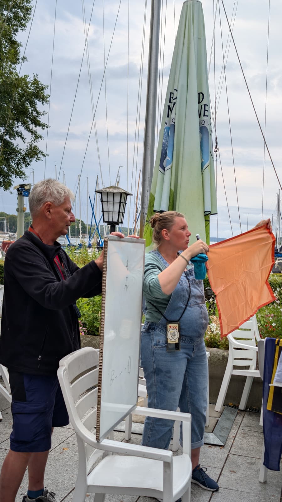 Zwei Helfer der Wettfahrtleitung halten auf der Terrasse des SC Gothia eine beschriebene Ergebnistafel und eine orangefarbene Flagge hoch, dahinter der Hafen