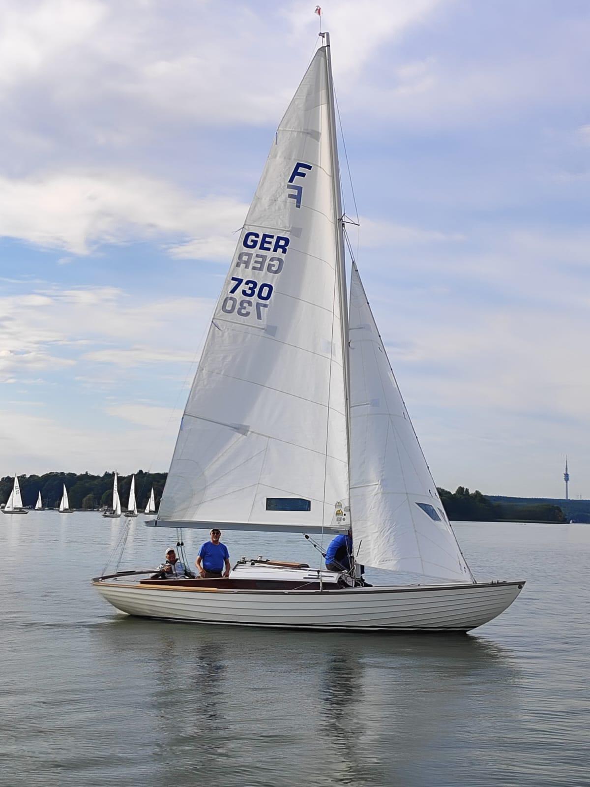 Nordisches Folkeboot GER 730 unter vollen Segeln bei leichtem Wind, drei Crewmitglieder an Bord, weitere Segelboote am Horizont