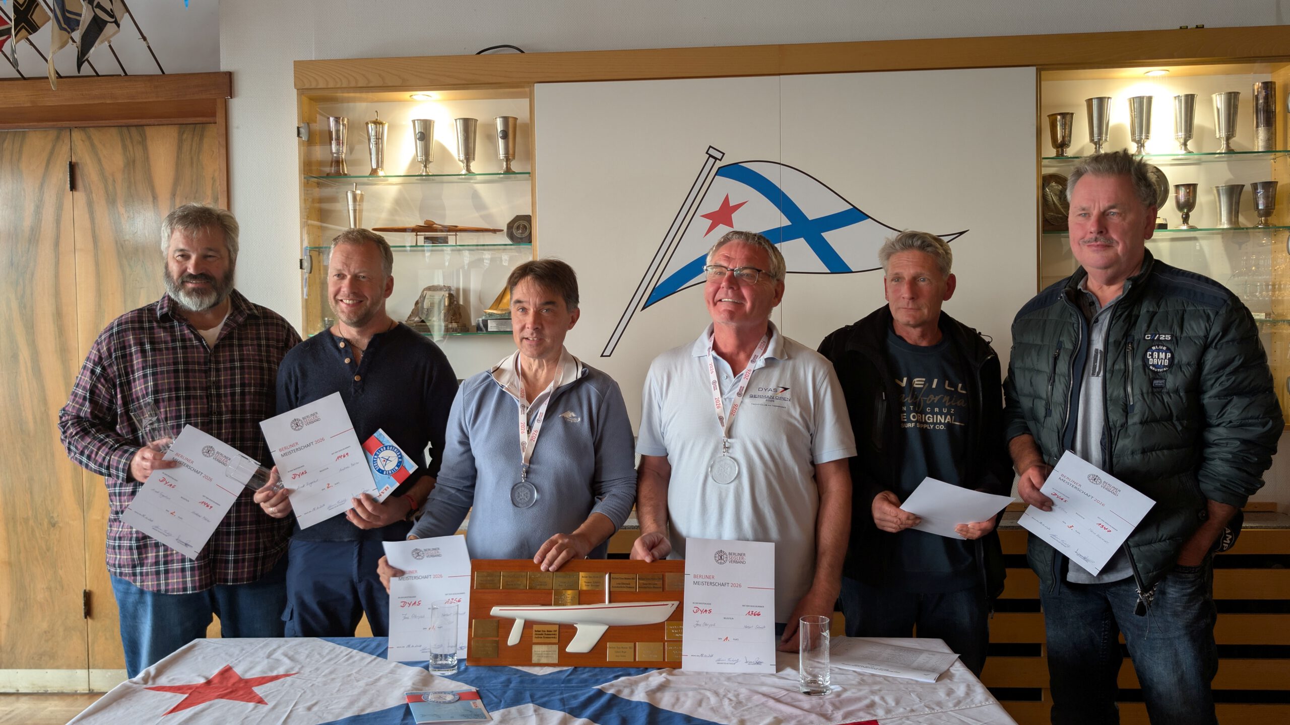 Sechs Segler mit Medaillen und Urkunden bei der Siegerehrung des Baumann-Preises im Clubhaus des SC Gothia, vor ihnen der Wanderpreis mit Dyas-Modell