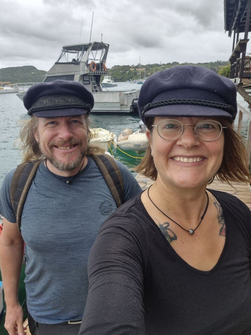 Henri Bergius und Susanna Huhtanen mit Segelmützen lächelnd auf einem Steg, im Hintergrund Boote in einer Bucht im Südpazifik